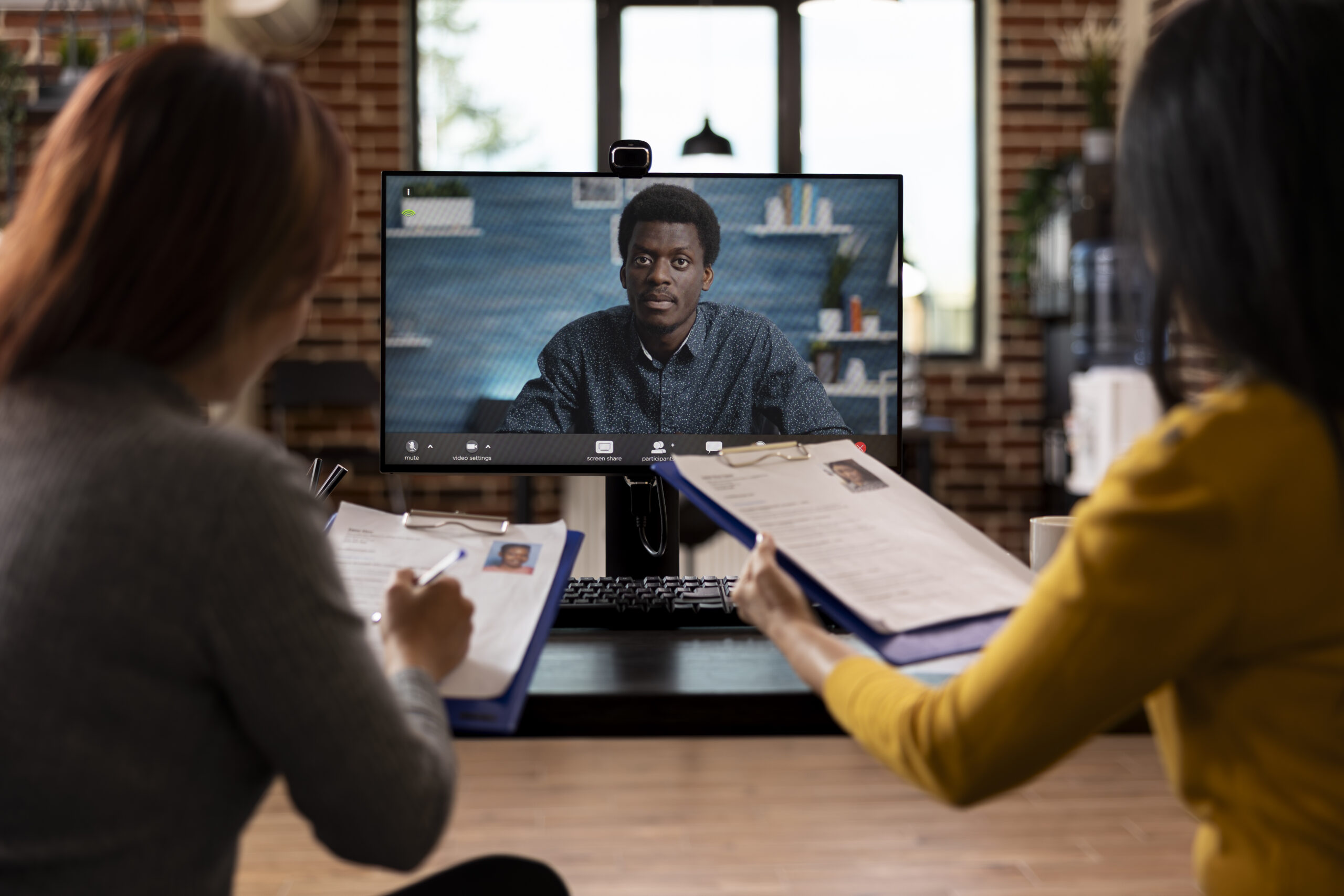 Female HR team conducts a virtual meeting with african american man, assessing CVs and discussing recruitment. Asian women holding clipboards engage in a video call with black manager on desktop pc.